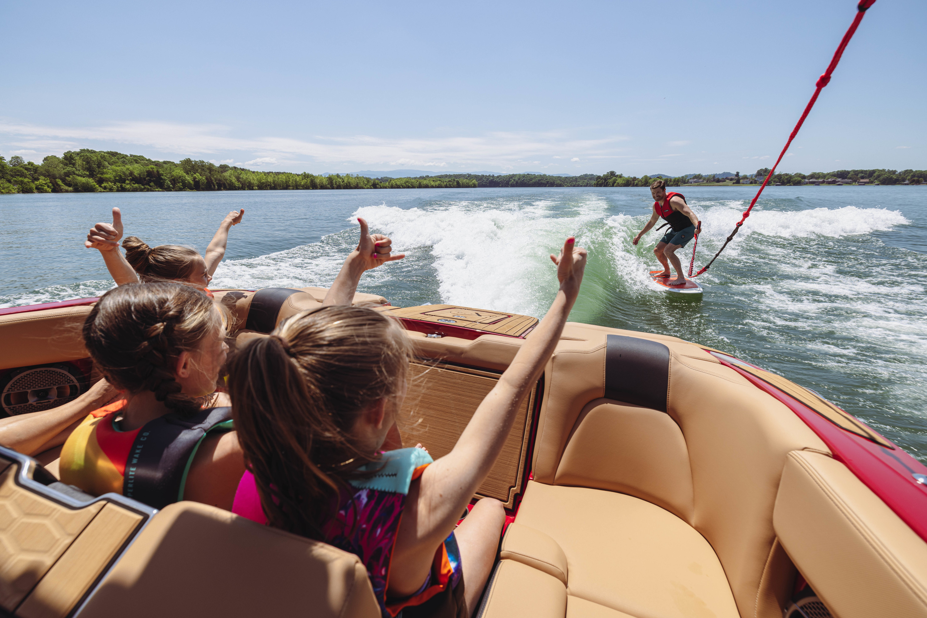 Two kids facing the wake on a MasterCraft Boat as a wakesurfer holds a surf rope.